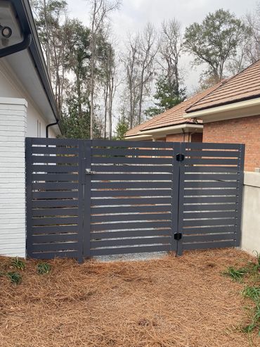 Modern black slatted gate next to a white house and brick wall.