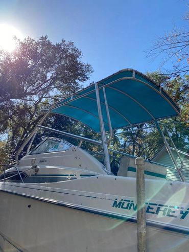 A Monterey boat with a blue canopy docked near trees on a sunny day.